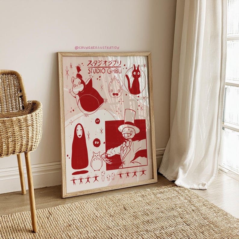 May include: A framed poster featuring red illustrations of Studio Ghibli characters on a pink background. The poster includes characters from various Ghibli films, with Japanese text at the top. The frame is made of light-colored wood.