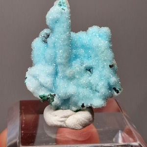 Op de afbeelding: Een levendig, lichtblauw mineraal specimen met een kristallijne structuur. Het mineraal heeft een textuurrijk oppervlak met kleine, sprankelende insluitsels. Donkere vlekken zijn verspreid over het oppervlak. Het mineraal staat op een witte basis, gemonteerd op een heldere displaystandaard.