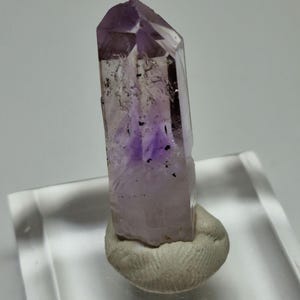 Può includere: Un cristallo di ametista trasparente con una punta e un interno viola scuro, montato su una piccola base chiara. Il cristallo presenta inclusioni visibili e una superficie sfaccettata lucida.
