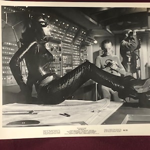 Könnte beinhalten: Schwarz-Weiß-Fotografie aus der Batman-Fernsehserie, die eine Frau in einem Catwoman-Kostüm zeigt, die auf einem Tisch sitzt. Im Hintergrund sind zwei Männer zu sehen. Das Set hat eine Retro-Futurismus-Ästhetik.