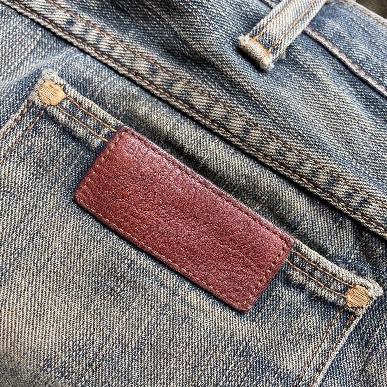 May include: Close-up of denim jeans with a brown leather label. The label is rectangular and stitched to the back pocket. The label has the text "BLUE BELLS" and a design. The jeans are faded blue.