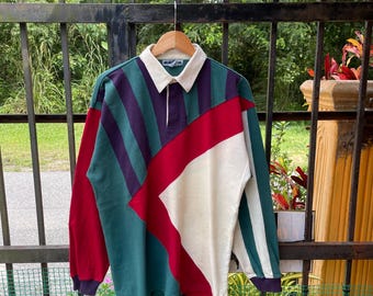 Può includere: Una maglia da rugby a maniche lunghe con colletto bianco e design a blocchi di colore. La maglia presenta pannelli verdi, viola, rossi e bianchi. Le maniche sono rosse e verdi con polsini viola. La maglia è appesa a una gruccia di legno.