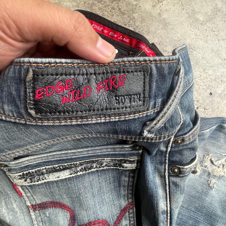 K&ouml;nnte beinhalten: Nahaufnahme einer blauen Jeans mit Used-Look. Die Jeans hat einen schwarzen Lederaufn&auml;her mit den rot gestickten Worten "EDGE WILD FIRE". Der Markenname "EDWIN" ist ebenfalls sichtbar. Die Jeans hat rote N&auml;hte und Metallnieten.
