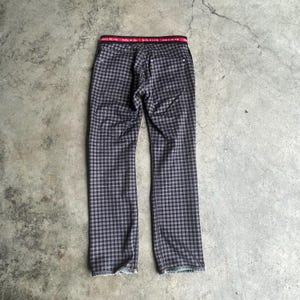 K&ouml;nnte beinhalten: Eine Hose mit schwarz-grauem Karomuster. Sie hat einen roten Bund mit wei&szlig;em Text. Die Hose liegt flach auf einer grauen Oberfl&auml;che. Das Muster ist im Vichy-Stil gehalten.