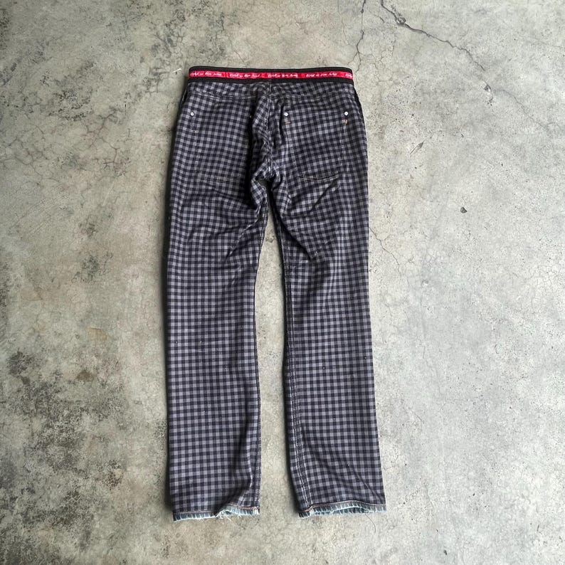 K&ouml;nnte beinhalten: Eine Hose mit schwarz-grauem Karomuster. Sie hat einen roten Bund mit wei&szlig;em Text. Die Hose liegt flach auf einer grauen Oberfl&auml;che. Das Muster ist im Vichy-Stil gehalten.