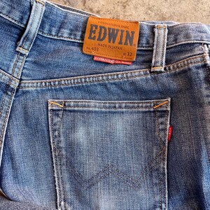 Pu&ograve; includere: Primo piano di jeans blu con tasca posteriore e toppa in pelle marrone. La toppa recita "EDWIN" e "MADE IN JAPAN". I jeans hanno un'etichetta rossa sulla tasca posteriore e sono taglia W32.