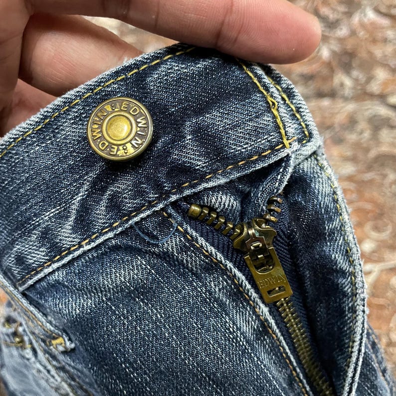 May include: A pair of dark blue denim jeans featuring a brass-toned metal button embossed with the text EDWIN and a matching brass-toned metal zipper pull also engraved with the brand name EDWIN. The fabric displays a classic indigo wash.