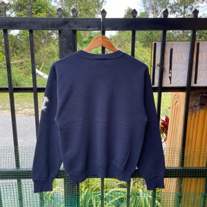 May include: Navy blue sweatshirt with a white letter graphic on the left sleeve. The sweatshirt is hanging on a wooden hanger against a metal gate. The background includes greenery and a glimpse of a building.