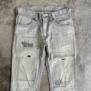 K&ouml;nnte beinhalten: Hellgraue Jeans aus Denim mit abgenutzten Patches und schwarzen Nahtdetails. Die Jeans haben ein klassisches F&uuml;nf-Taschen-Design und einen Knopfverschluss. Die Patches haben ein einzigartiges grafisches Design.