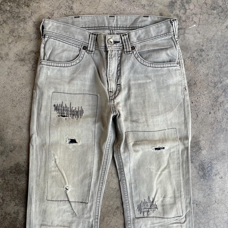 K&ouml;nnte beinhalten: Hellgraue Jeans aus Denim mit abgenutzten Patches und schwarzen Nahtdetails. Die Jeans haben ein klassisches F&uuml;nf-Taschen-Design und einen Knopfverschluss. Die Patches haben ein einzigartiges grafisches Design.
