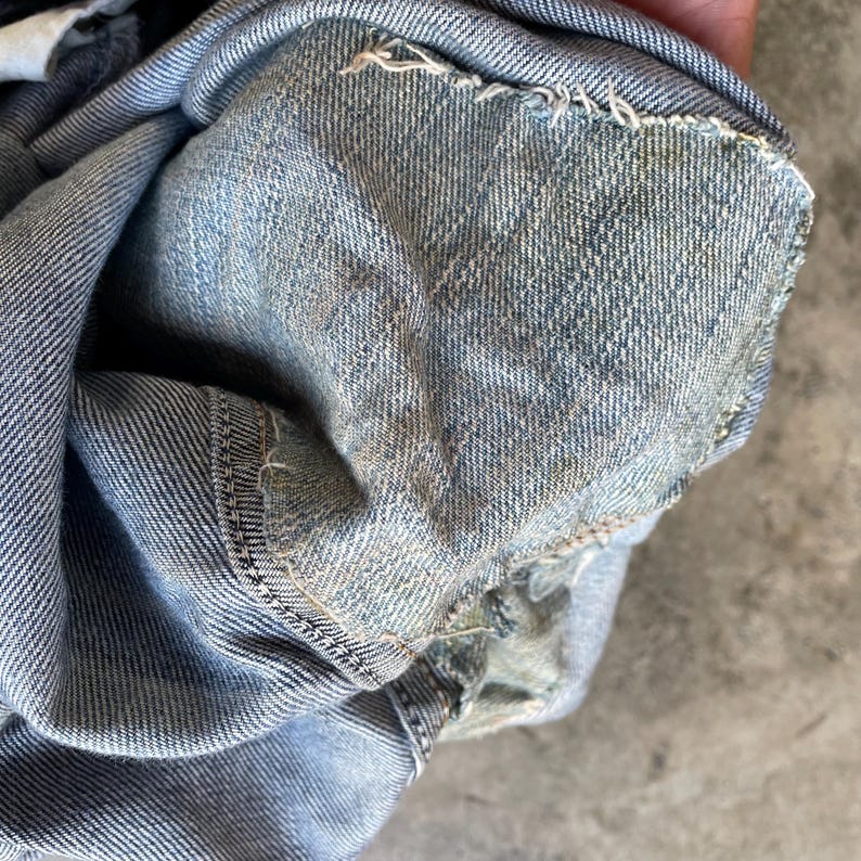 May include: Close-up of light blue denim fabric, displaying the texture and stitching. The denim has a worn look with frayed edges, suggesting a vintage or distressed style. The fabric is folded, revealing the inside.