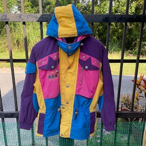 Könnte beinhalten: Eine mehrfarbige Skijacke in den Farben Gelb, Pink, Blau und Lila. Die Jacke hat eine Kapuze und einen Reißverschluss. Die Jacke hat ein Logo mit der Aufschrift "Rockland" auf der Vorderseite.