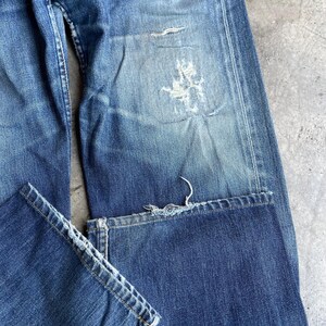 May include: Close-up of distressed blue denim jeans. The jeans show signs of wear and tear, with faded areas and a large rip on the right leg. The hem of the jeans is frayed, adding to the vintage aesthetic. The denim has a classic, worn look.
