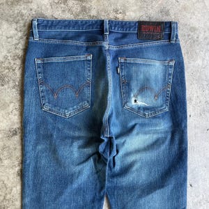 Op de afbeelding: Close-up van de achterkant van een blauwe spijkerbroek. De jeans heeft twee achterzakken met een kenmerkend rood stikselontwerp. Een leren patch met de merknaam "EDWIN" is zichtbaar bij de tailleband. De denim heeft een versleten, vervaagde uitstraling.