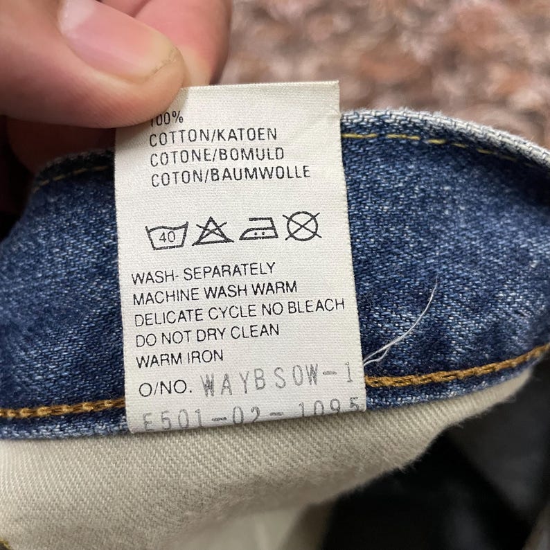 May include: A white fabric care label sewn into blue denim fabric. The label reads 100% COTTON/KATOEN COTONE/BOMULD COTON/BAUMWOLLE, includes laundry symbols, and instructions: WASH SEPARATELY, MACHINE WASH WARM, DELICATE CYCLE NO BLEACH, DO NOT DRY CLEAN, WARM IRON.