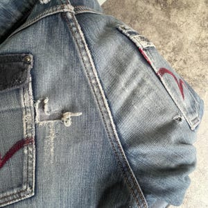 K&ouml;nnte beinhalten: Nahaufnahme einer hellblauen Jeansjacke mit Used-Details. Zu den Merkmalen geh&ouml;ren ein rotes Stickdesign auf der Tasche und ein zerrissener Bereich in der N&auml;he der Tasche. Wei&szlig;e N&auml;hte akzentuieren die N&auml;hte. Die Jacke hat einen Vintage-Look.