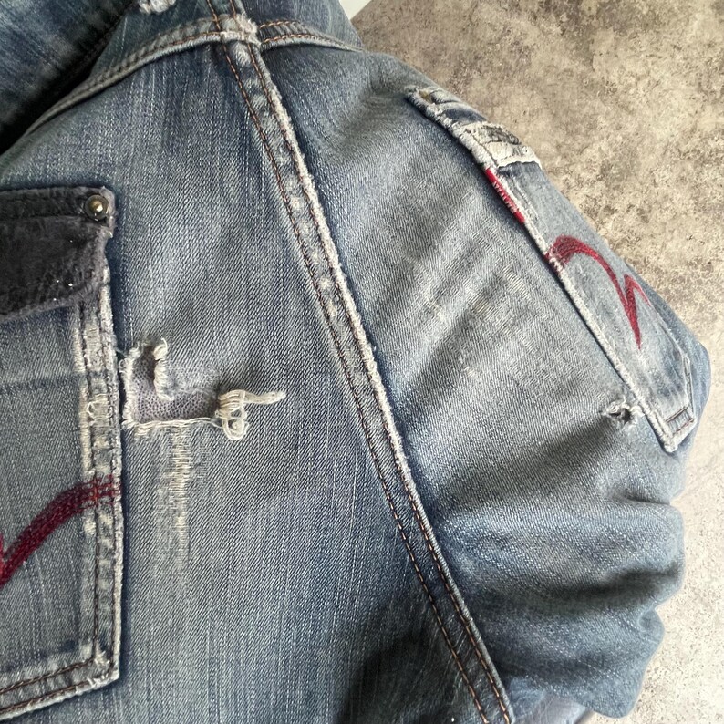 K&ouml;nnte beinhalten: Nahaufnahme einer hellblauen Jeansjacke mit Used-Details. Zu den Merkmalen geh&ouml;ren ein rotes Stickdesign auf der Tasche und ein zerrissener Bereich in der N&auml;he der Tasche. Wei&szlig;e N&auml;hte akzentuieren die N&auml;hte. Die Jacke hat einen Vintage-Look.