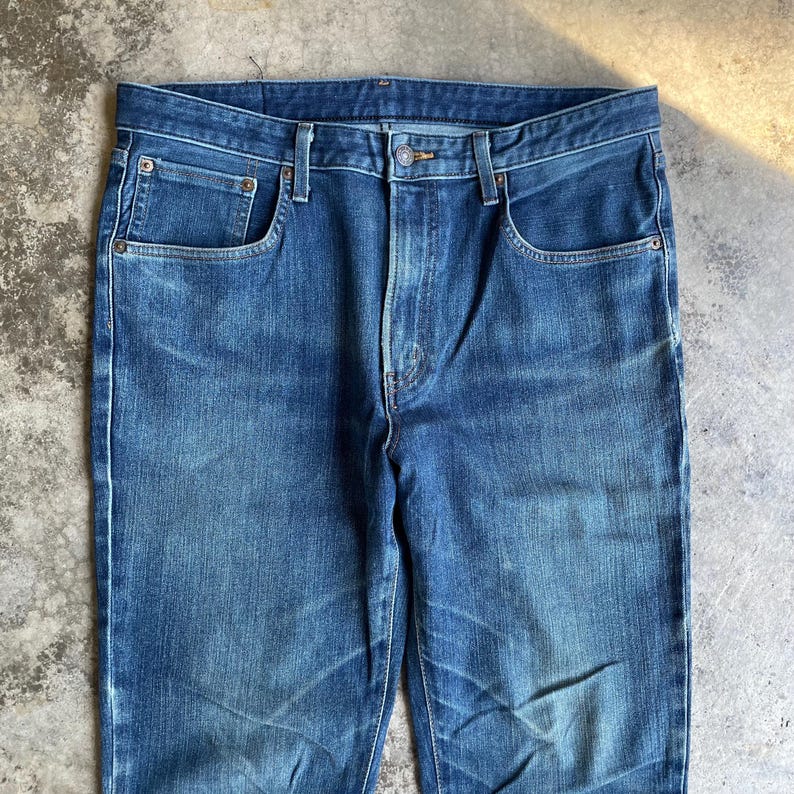 Op de afbeelding: Close-up van een blauwe spijkerbroek. De jeans heeft een klassiek ontwerp met vijf zakken, een knoop- en ritssluiting en een vervaagde wassing. De denimstof vertoont slijtage, wat bijdraagt aan de vintage esthetiek.