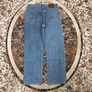 May include: Light blue denim jeans featuring a straight-leg cut, a tan leather patch on the back waistband, and two back pockets with a signature W-shaped stitching pattern. The jeans include a small red tab on the right back pocket.