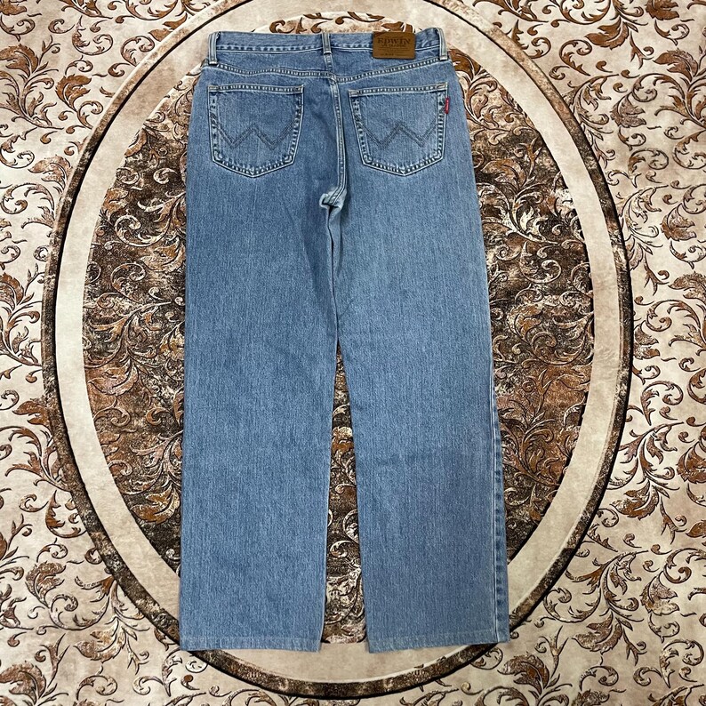 May include: Light blue denim jeans featuring a straight-leg cut, a tan leather patch on the back waistband, and two back pockets with a signature W-shaped stitching pattern. The jeans include a small red tab on the right back pocket.