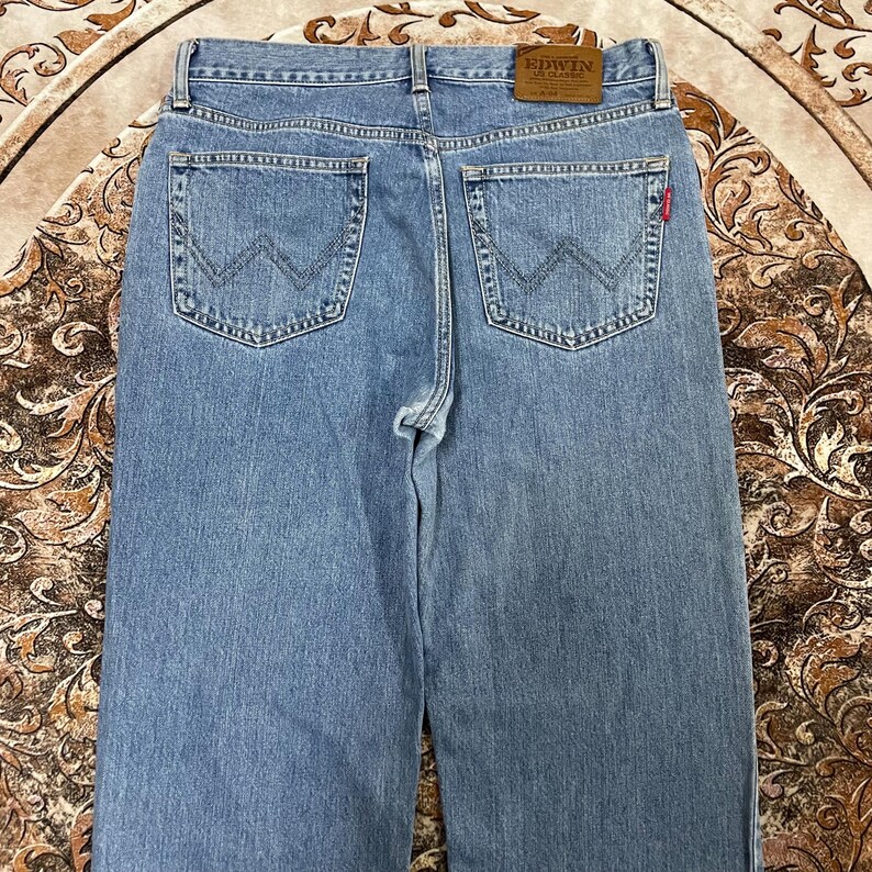 May include: Light blue denim jeans featuring a classic five-pocket design with a high waist. The back pockets display a signature W-shaped stitching pattern. A brown leather patch with the text EDWIN US CLASSIC is attached to the waistband.
