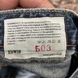 May include: A blue denim fabric waistband featuring a rectangular white interior label with the text EDWIN 503, including care instructions and a paragraph describing the cotton quality, with the brand name EDWIN and website URL printed.