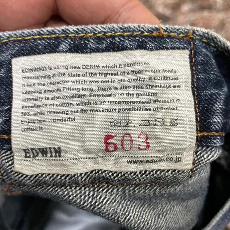 May include: A blue denim fabric waistband featuring a rectangular white interior label with the text EDWIN 503, including care instructions and a paragraph describing the cotton quality, with the brand name EDWIN and website URL printed.