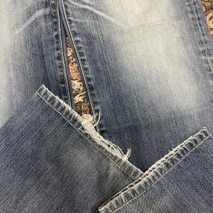 May include: A pair of blue denim jeans featuring a faded wash and a distressed, frayed hemline at the bottom of the pant legs. The fabric displays a classic twill texture with visible stitching along the seams and a worn-in appearance throughout.