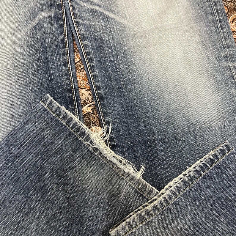 May include: A pair of blue denim jeans featuring a faded wash and a distressed, frayed hemline at the bottom of the pant legs. The fabric displays a classic twill texture with visible stitching along the seams and a worn-in appearance throughout.