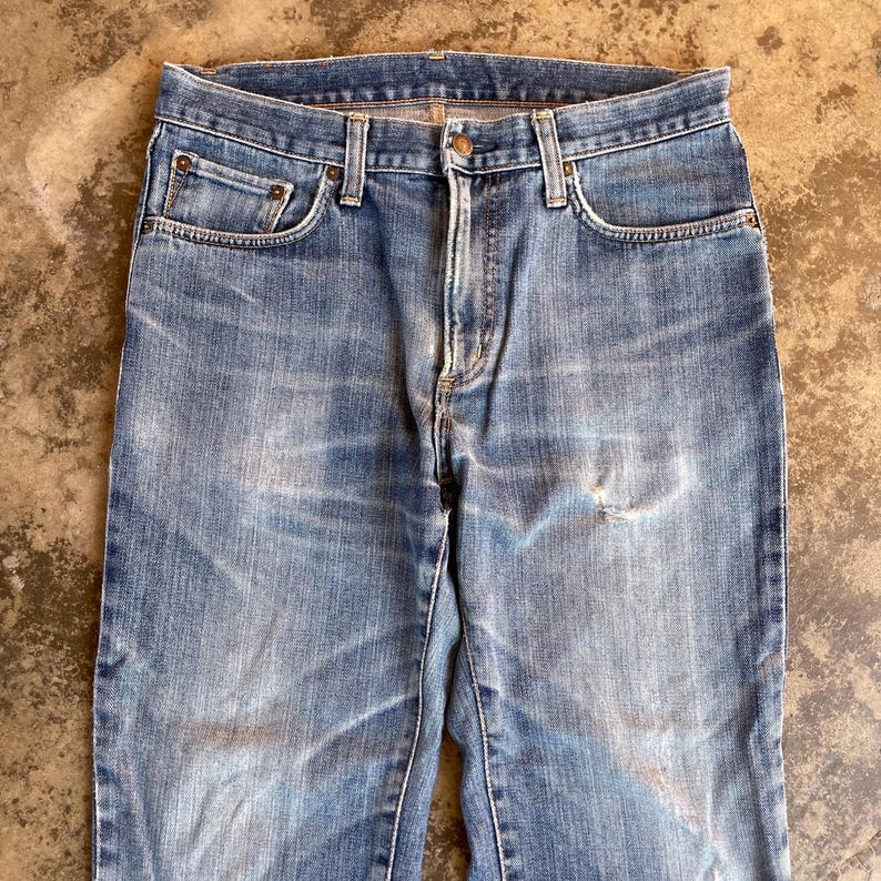 Pu&ograve; includere: Jeans di denim blu chiaro con un classico design a cinque tasche. I jeans mostrano segni di usura con aree sbiadite e un piccolo strappo sulla gamba destra. I jeans hanno una chiusura con bottone e cerniera e passanti per cintura.