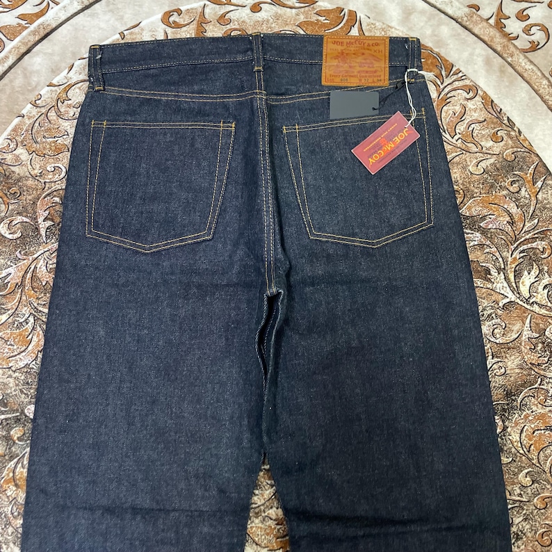 May include: Dark blue denim jeans with yellow stitching. The back of the jeans features two square pockets and a leather patch at the waistband. A red tag with the text "JOE McCOY" is attached.