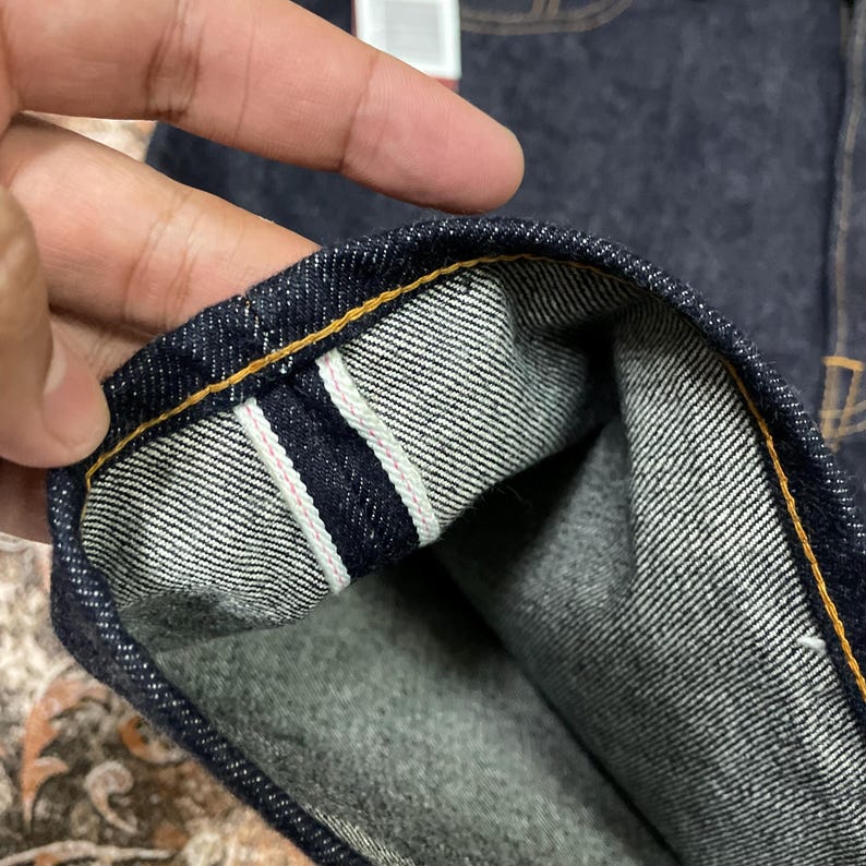 May include: Close-up of the hem of dark blue denim jeans. The hem is turned up, revealing a white and pink stripe detail. The stitching is a contrasting yellow color.
