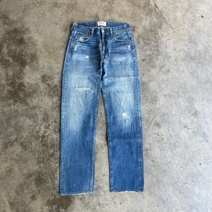 Puede incluir: Vaqueros de mezclilla azul descolorido con detalles desgastados, incluyendo una rodilla rasgada y parches desgastados. Los vaqueros tienen un diseño clásico de cinco bolsillos y bragueta con botones. El aspecto general es vintage e informal.