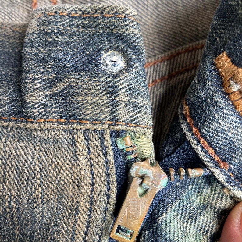 May include: Close-up of a worn denim jeans zipper. The zipper is a tarnished brass color with the word "UNIVERSAL" stamped on it. The denim is faded blue with visible wear and tear, and orange stitching.