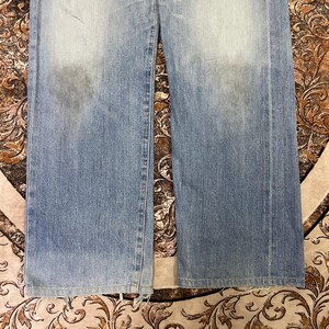 May include: Light blue denim jeans with a faded wash and a straight leg cut. The jeans show signs of wear and tear, with some fraying at the bottom hem. The fabric has a slightly distressed appearance.