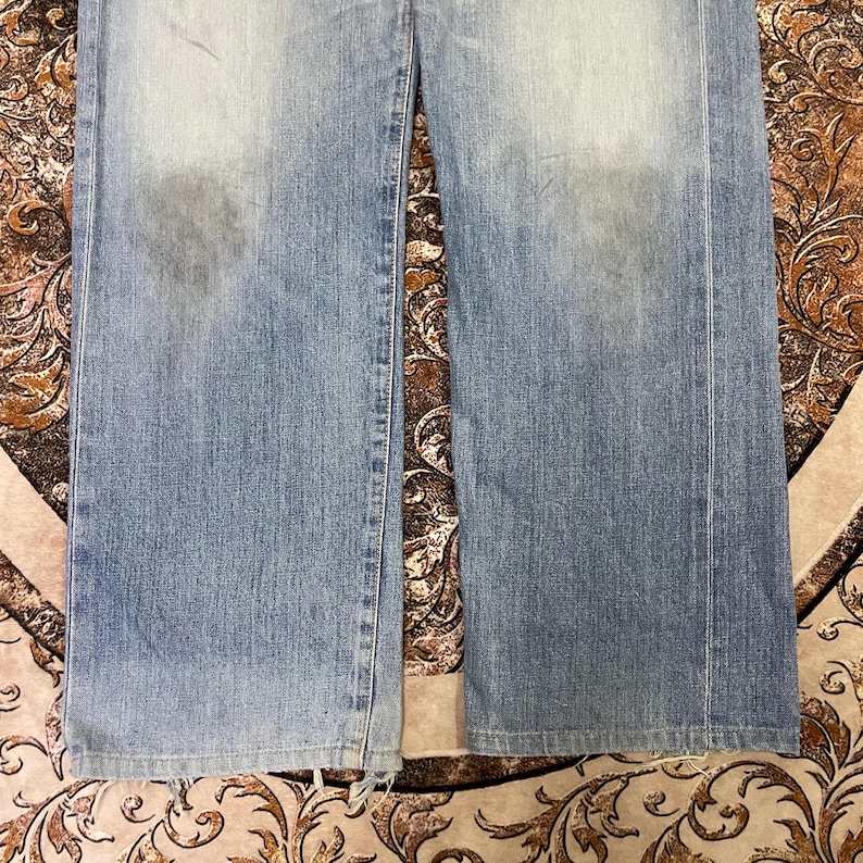 May include: Light blue denim jeans with a faded wash and a straight leg cut. The jeans show signs of wear and tear, with some fraying at the bottom hem. The fabric has a slightly distressed appearance.