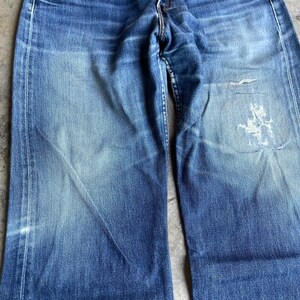 May include: Close-up of faded blue denim jeans. The jeans show wear and tear, with visible fading and distressed areas. The fabric has a textured appearance, with lighter shades of blue creating a worn look. The jeans have a classic five-pocket design.