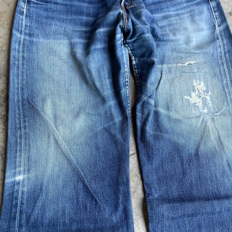 May include: Close-up of faded blue denim jeans. The jeans show wear and tear, with visible fading and distressed areas. The fabric has a textured appearance, with lighter shades of blue creating a worn look. The jeans have a classic five-pocket design.