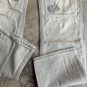 K&ouml;nnte beinhalten: Hellgraue Jeans mit schwarzen N&auml;hten und einem Design mit abgenutzter Tasche. Die Jeans sind gefaltet, wodurch der Saum und die Textur des Stoffes sichtbar werden. Der Gesamteindruck ist l&auml;ssig und abgenutzt.