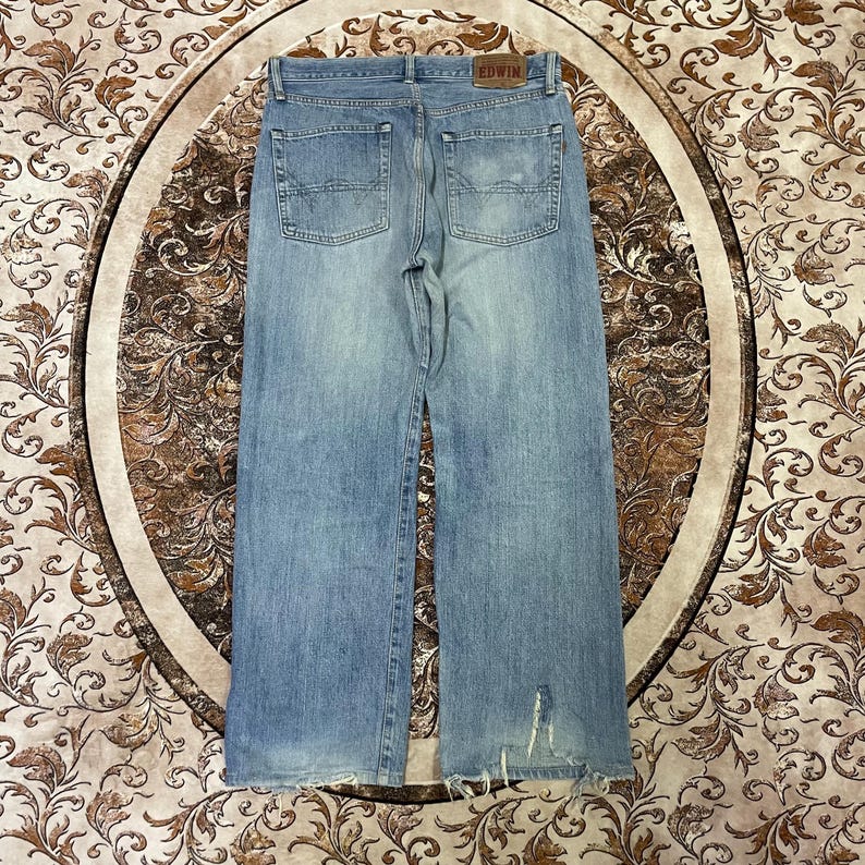 May include: Light blue denim jeans with a wide leg cut. The jeans have a faded wash and distressed details at the hem. The back pockets are visible, and a brown leather patch with the brand name is at the waistband.