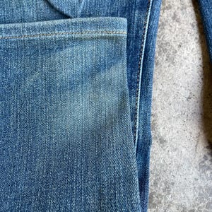 Op de afbeelding: Close-up van een blauwe spijkerbroek, met details van de zak en naden. De denim heeft een vervaagde uitstraling met zichtbare textuur en stiksels. De jeans hebben een medium wassing.