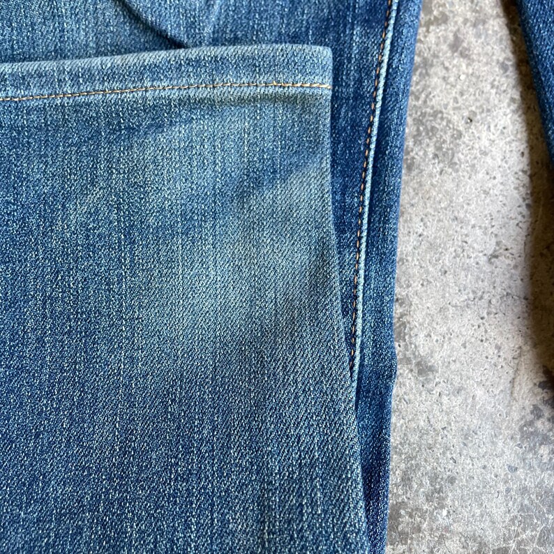 Op de afbeelding: Close-up van een blauwe spijkerbroek, met details van de zak en naden. De denim heeft een vervaagde uitstraling met zichtbare textuur en stiksels. De jeans hebben een medium wassing.
