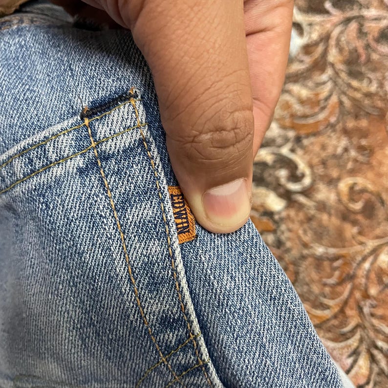May include: Close-up of light blue denim fabric with orange stitching and a small orange Edwin label. The denim has a classic, worn look, with visible seams and a pocket detail. The fabric is held by a hand.