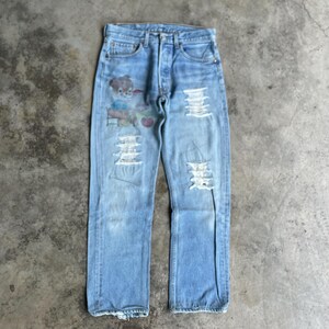 Puede incluir: Vaqueros de mezclilla azul claro con detalles desgastados y un diseño de oso de peluche pintado en la parte delantera. Los vaqueros tienen múltiples secciones rasgadas y un estilo clásico de cinco bolsillos. Prenda de inspiración vintage.