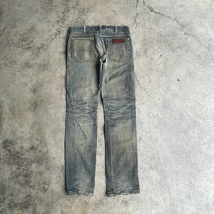 May include: Faded blue denim jeans with a worn appearance. The jeans have a classic five-pocket design, a brown leather patch, and distressed detailing on the legs. The jeans are displayed on a gray concrete surface.