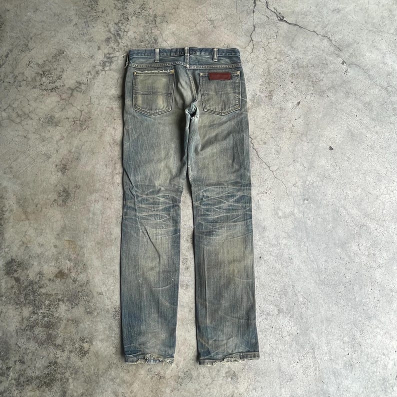May include: Faded blue denim jeans with a worn appearance. The jeans have a classic five-pocket design, a brown leather patch, and distressed detailing on the legs. The jeans are displayed on a gray concrete surface.