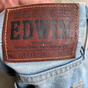 May include: Close-up of a light blue denim jeans label. The brown leather label features the brand name "EDWIN" in bold black letters. The label also includes text about the jeans and the number "505ZXX".