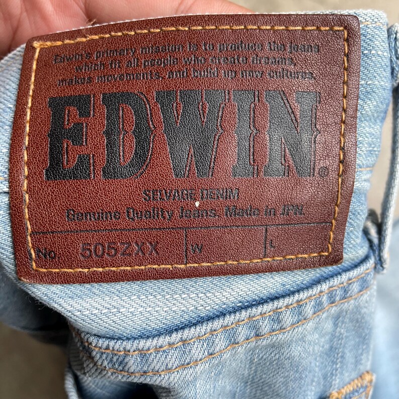 May include: Close-up of a light blue denim jeans label. The brown leather label features the brand name "EDWIN" in bold black letters. The label also includes text about the jeans and the number "505ZXX".