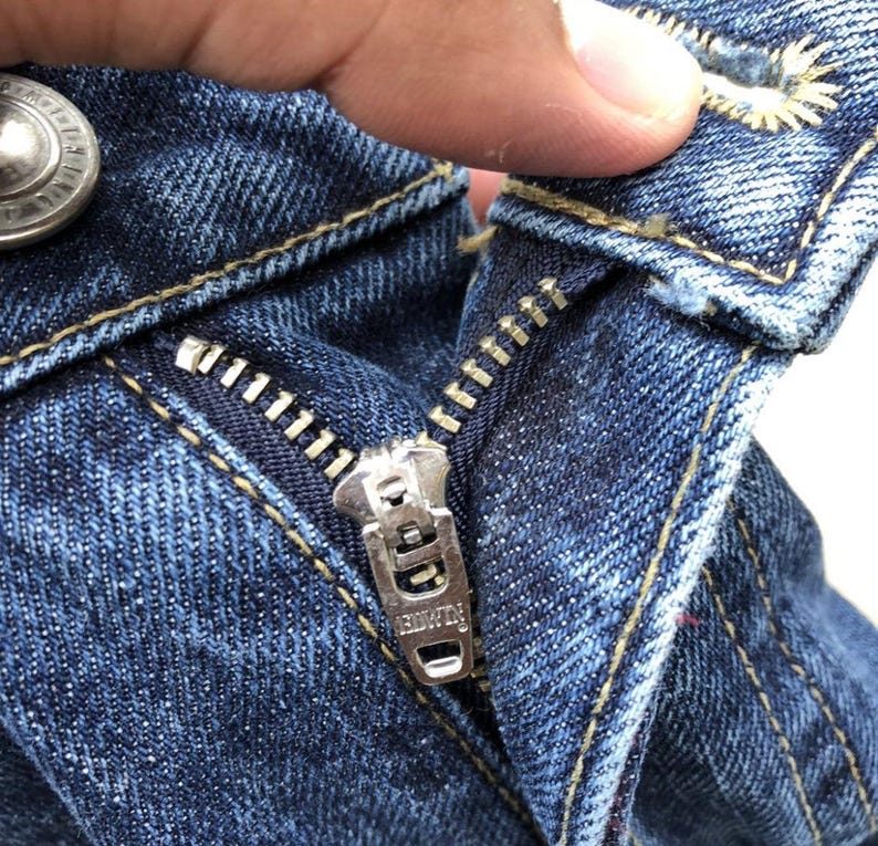 Op de afbeelding: Close-up van een blauwe denim ritsvlieg met een metalen rits-trekker waarop "R.I.V.E.T.T.E." staat.