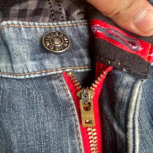 K&ouml;nnte beinhalten: Nahaufnahme einer blauen Jeans mit rotem Rei&szlig;verschluss und Metallknopf. Der goldfarbene Rei&szlig;verschluss tr&auml;gt den Markennamen "EDWIN". Der Knopf tr&auml;gt ebenfalls den Markennamen "EDWIN". Die Jeans hat innen ein Karomuster.